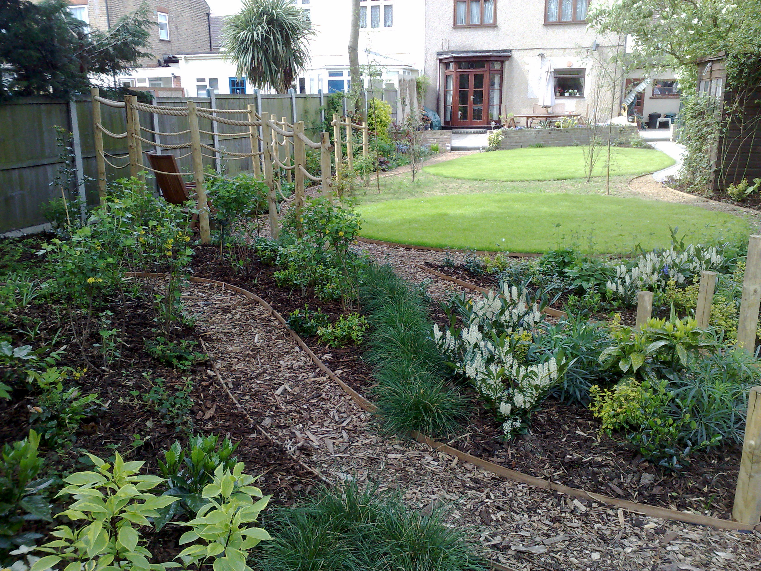 View of garden looking back to the house across the winding shady walk path with shrubs and seating area in midgorund